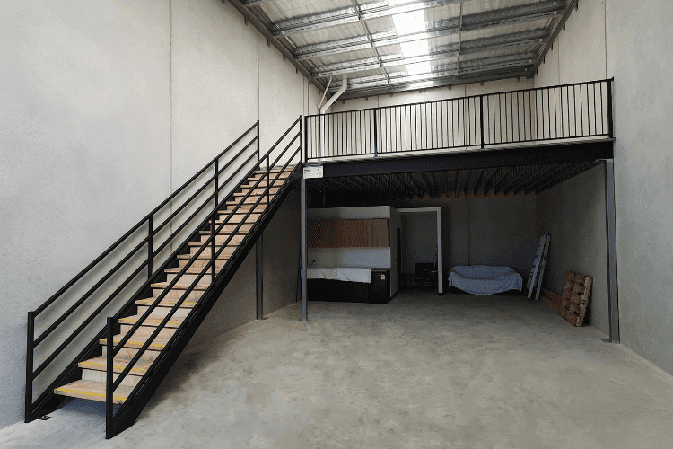 Compact warehouse interior featuring a black steel mezzanine, stairs, and ground-level workspace.