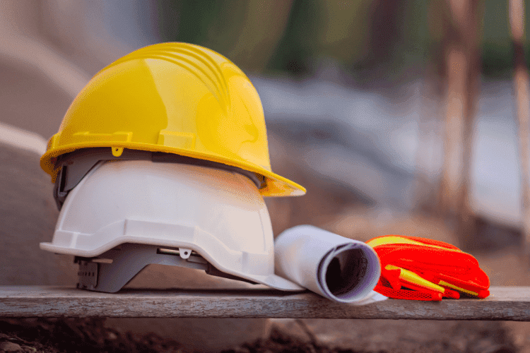 Yellow and white safety helmets placed beside gloves and blueprints on a wooden surface at a construction site.