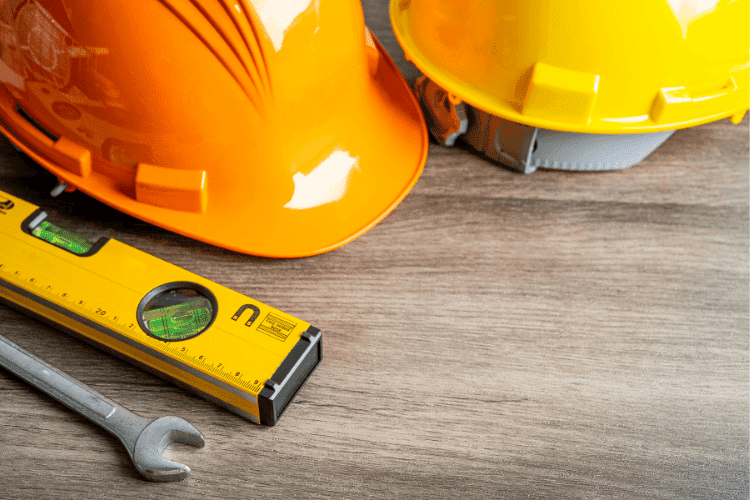 Orange and yellow hard hats placed beside a wrench and spirit level on a wooden surface.