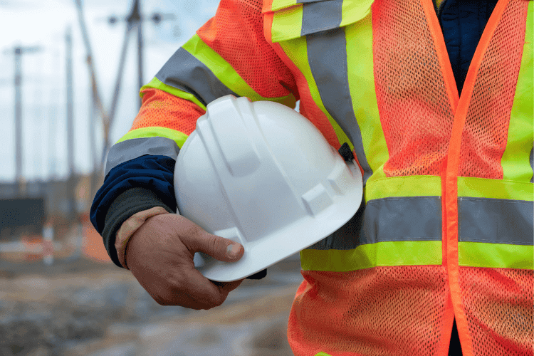 Construction worker in high-visibility vest holding a white safety helmet at a worksite.