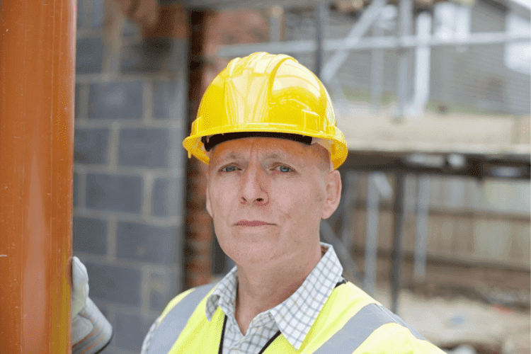 Construction supervisor wearing a yellow hard hat and reflective vest standing at a building site.