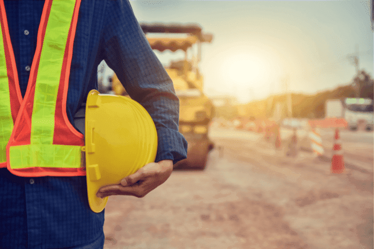 Construction worker wearing a reflective safety vest holding a yellow hard hat at a roadwork site during sunset.