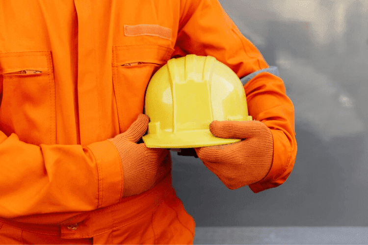 Worker wearing orange coveralls and gloves holding a yellow safety helmet against a gray background.