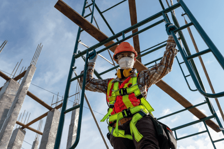 Construction worker in reflective vest and safety harness standing on scaffolding under a clear sky.