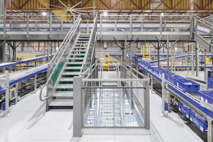 Elevated mezzanine structure with metal stairs and blue conveyor systems inside a distribution center.