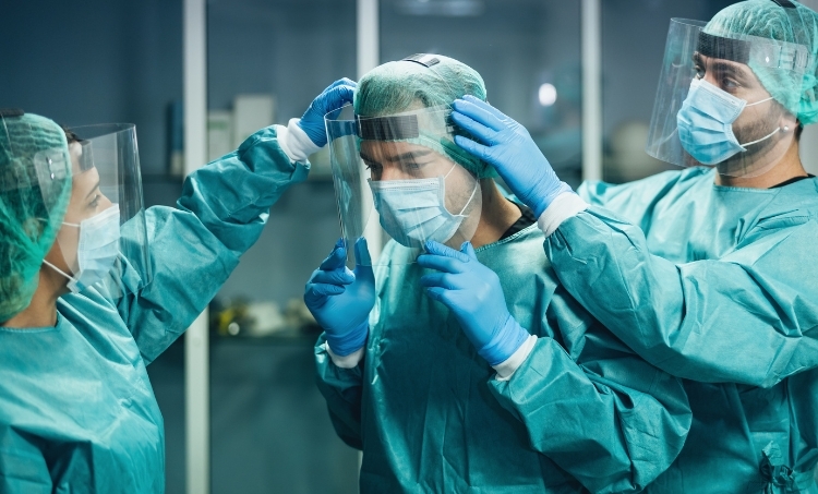 Three medical professionals in full protective gear help each other adjust face shields and masks, demonstrating proper PPE use and teamwork in a clinical environment.
