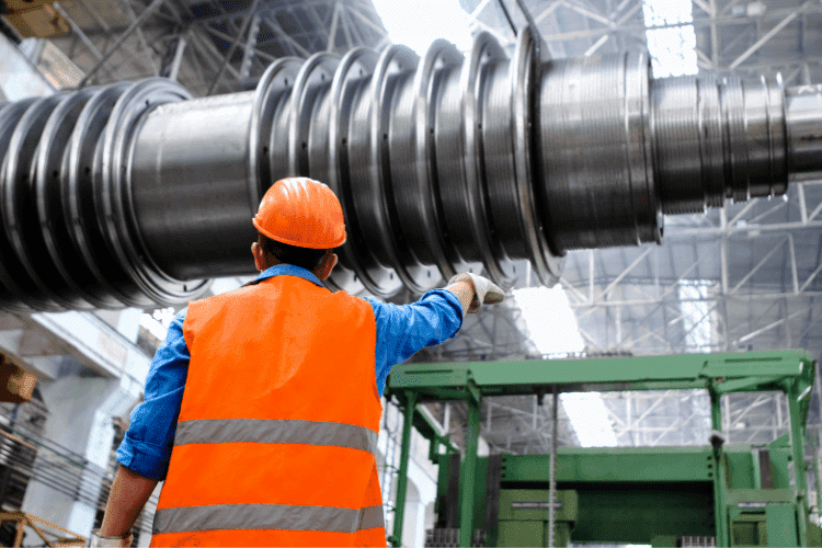 Industrial worker wearing orange vest and helmet inspecting large metal machinery in a factory.