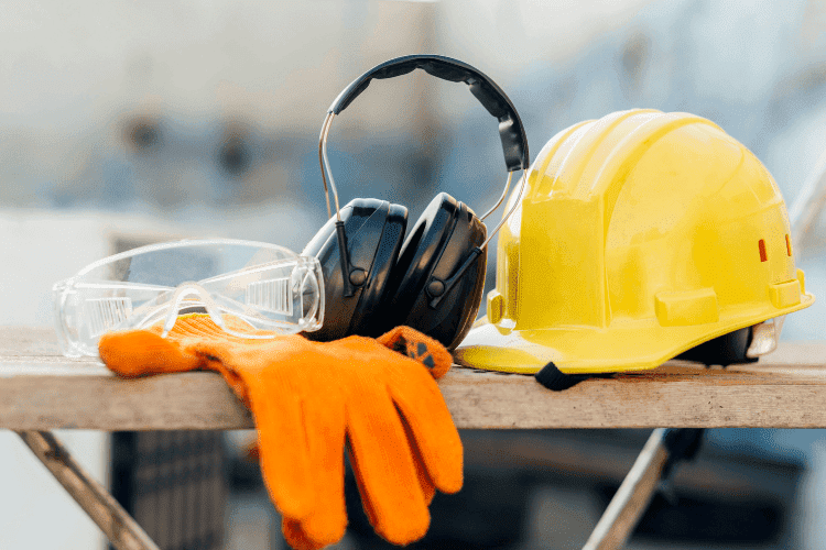 Set of construction safety gear including yellow hard hat, orange gloves, protective glasses, and earmuffs on a wooden plank.