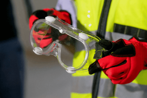 A close-up view of a worker wearing red and black safety gloves and a high visibility vest while holding a pair of clear protective safety goggles. The image highlights essential PPE used in industrial and construction environments.