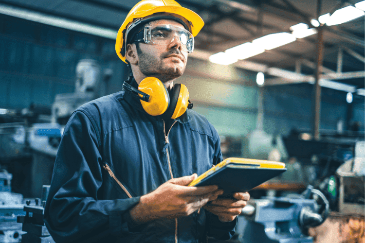 Industrial worker wearing yellow hard hat, protective glasses, and ear defenders while holding a tablet.