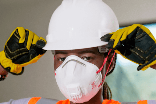 A construction worker wearing a white hard hat, yellow protective gloves, and a fitted respirator mask adjusts their helmet. The close up shot highlights essential personal protective equipment used on active job sites to reduce exposure to hazards.