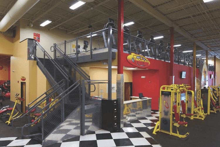 Fitness center with mezzanine floor, cardio equipment, and yellow gym machines on black-and-white checkered flooring.