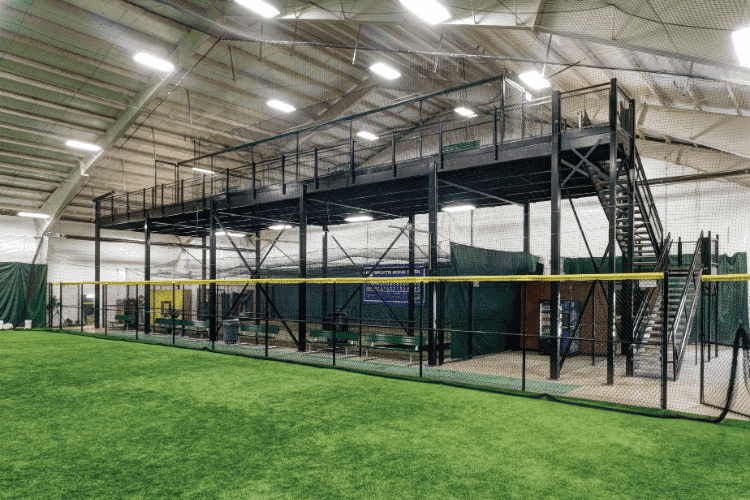 Indoor training facility with artificial turf and elevated mezzanine seating area behind safety nets.