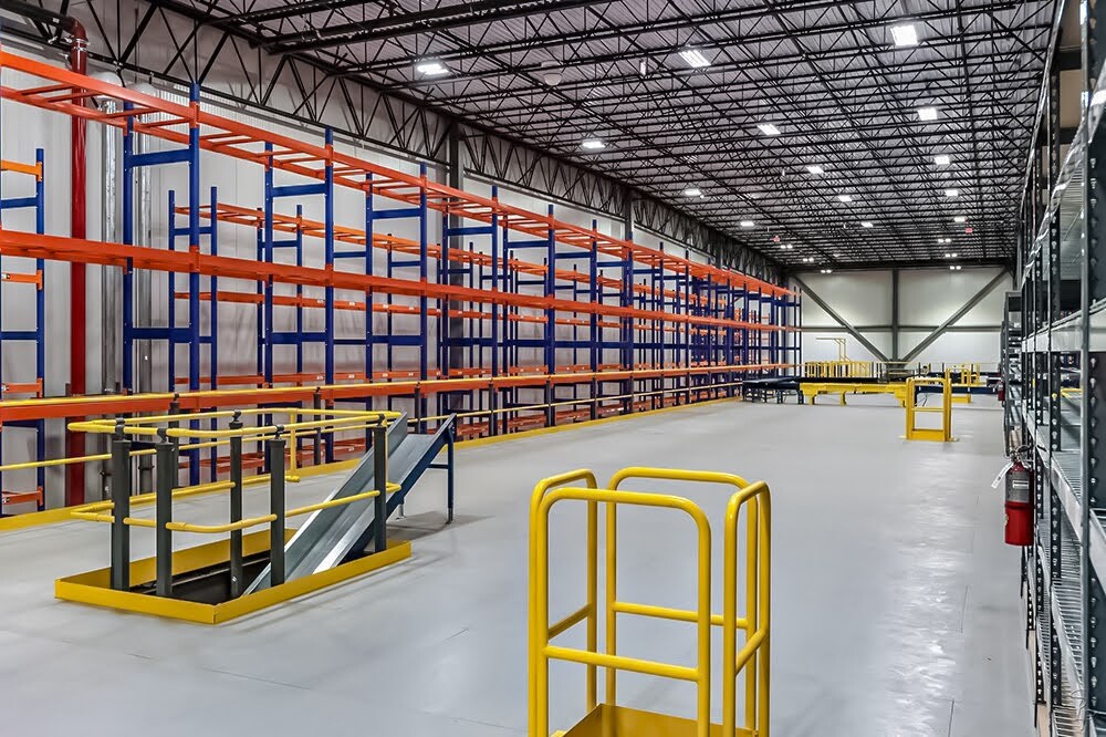 Large industrial warehouse with empty steel mezzanine floor, safety railings, and orange-blue pallet racking.
