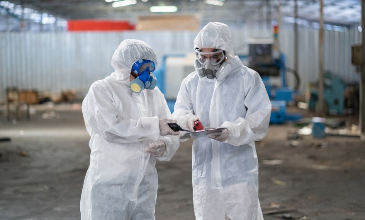 Two workers wearing full-body protective suits and respirators review data on a clipboard while performing a safety inspection inside an industrial facility.