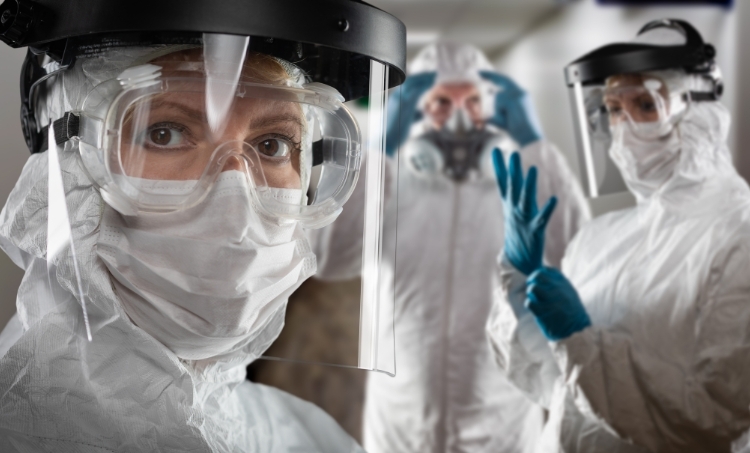 A close view of healthcare workers wearing full PPE, including face shields, goggles, masks, gloves, and protective suits. The foreground features a worker looking directly ahead, while others prepare equipment in the background, demonstrating strict safety protocols in a high-risk medical environment.