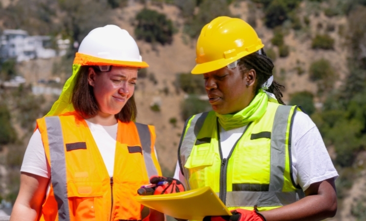 Two construction workers wearing hard hats, high visibility vests, and gloves review documents outdoors. Their protective gear highlights proper PPE use during site planning and safety discussions.