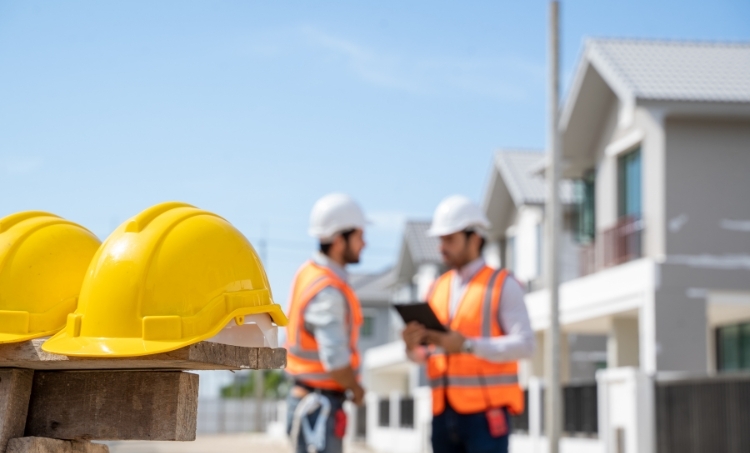 Two yellow hard hats sit on a wooden surface at a residential construction site while two workers in safety vests and helmets discuss plans in the background, highlighting the importance of PPE on active job sites.
