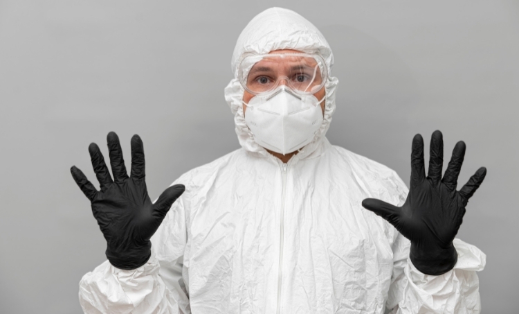 A person wearing full body protective clothing, including a white coverall suit, clear safety goggles, a white filtering facepiece respirator, and black protective gloves. The individual is facing the camera with both hands raised, illustrating the use of personal protective equipment in hazardous environments.