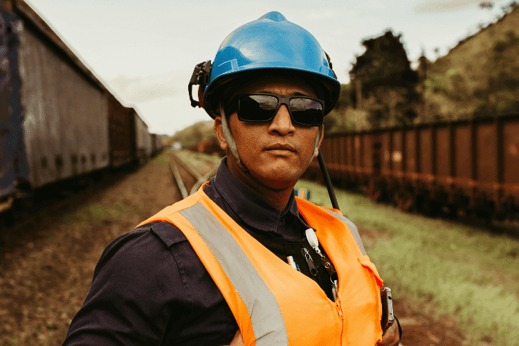 Railway worker in an orange vest and blue hard hat standing between train tracks wearing safety glasses.