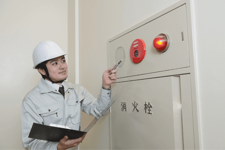 Safety inspector in white helmet checking a fire alarm panel and recording notes on clipboard.