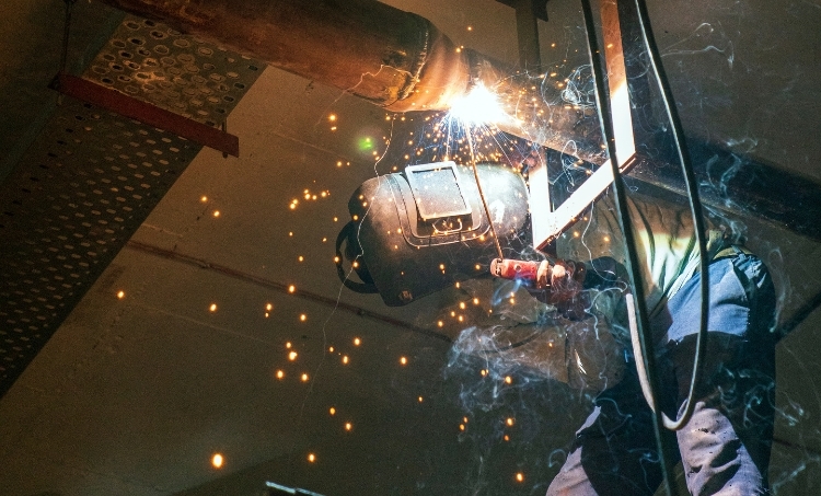 A worker welds overhead in a dim industrial setting, protected by a welding helmet, gloves, and flame-resistant clothing. Bright sparks and smoke surround the work area, highlighting the need for proper PPE during high heat and metalwork tasks.