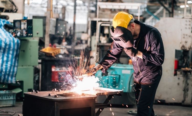 A person wearing protective gear welds metal on a workbench inside an industrial workshop. The individual is equipped with a hard hat and a welding helmet, with sparks visible as they work. The surrounding area includes machinery, tools, and workshop equipment.