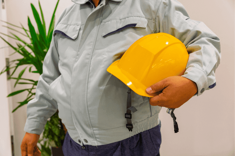 Worker in a light grey uniform holding a yellow safety helmet while standing inside a facility.