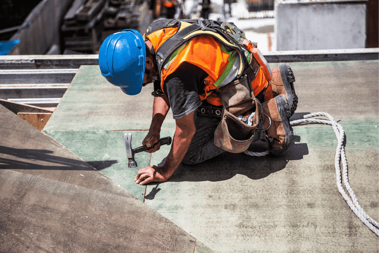 Construction worker in a blue hard hat and safety harness using a hammer while kneeling on a rooftop.