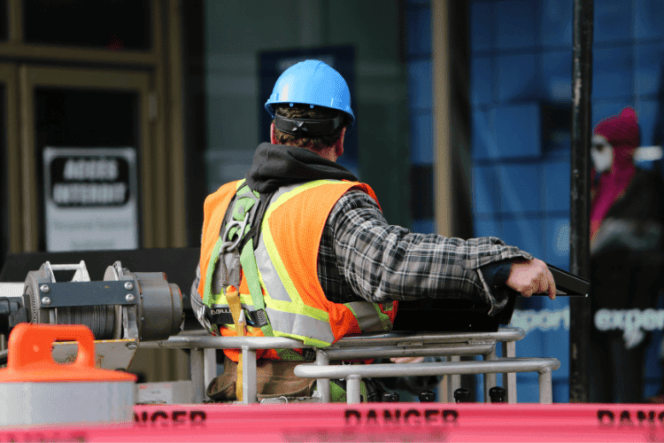 Construction worker wearing safety harness and blue helmet operating from an elevated lift behind danger tape.