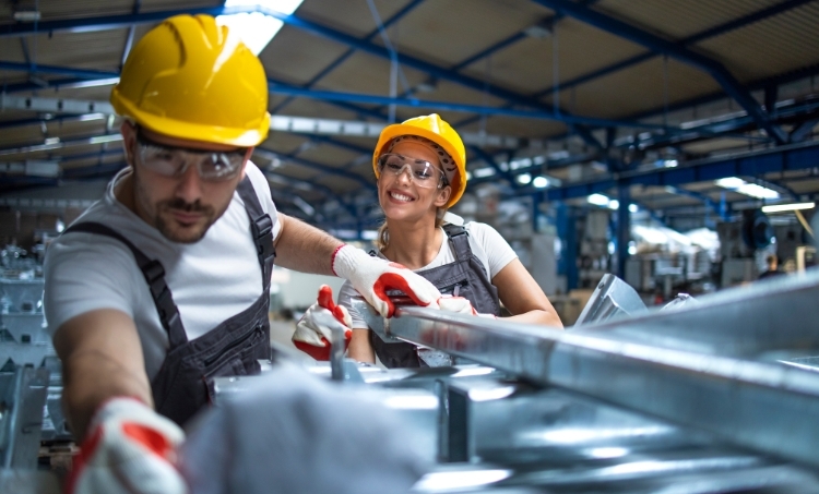 Two people wearing personal protective equipment work together in an industrial facility. Both are equipped with hard hats, safety glasses, gloves, and work overalls. One is operating equipment in the foreground while the other stands beside them smiling. The environment includes metal structures and machinery inside a large, well lit workshop.