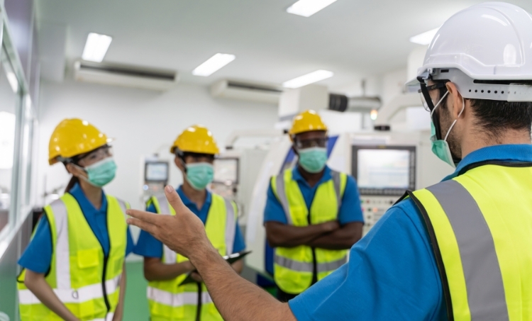 A group of workers wearing yellow hard hats, face masks, and high visibility safety vests listens to a supervisor during a safety briefing inside an industrial facility. The supervisor, also in protective gear, gestures while speaking. The setting includes bright overhead lighting and manufacturing equipment in the background.